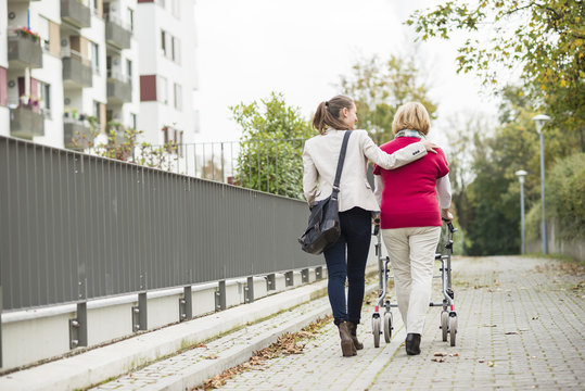 Adult granddaughter assisting her grandmother walking with wheeled walker, back view