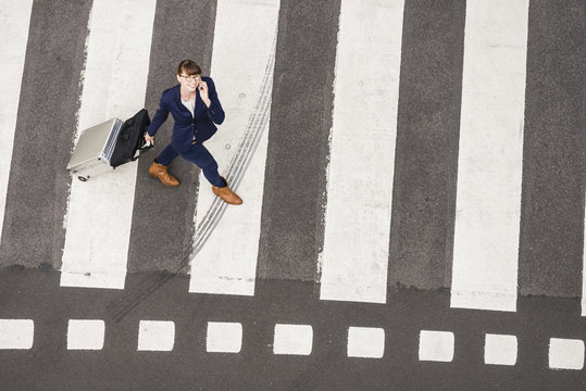 Germany, businesswoman with rolling suitcase walking on zebra crossing, view from above