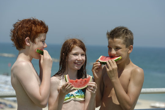 Italy, girl and two teenage boys eating watermelon slices at the beach