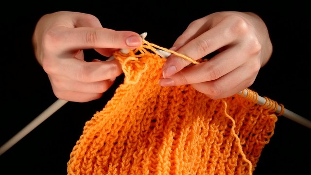 Woman sits and knits orange scarf, large knit on black