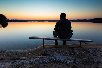 Man sitting on bench on lake shore at sunset