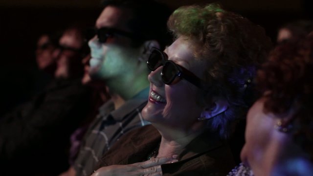 Mature woman watching a movie and wearing 3D glasses.  Focus on her with a small dolly move and projections on her face.