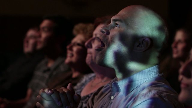 Young man has various reactions while watching a movie with a group of friends.  Ends with everyone clapping.  Focus on him with a small dolly move and projections on his face.