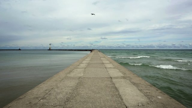 Concrete pier with calm water on one side and waves breaking on the other.  Lighthouse can be seen at the end of the harbor.  Recorded on Lake Michigan at Frankfort, Michigan.