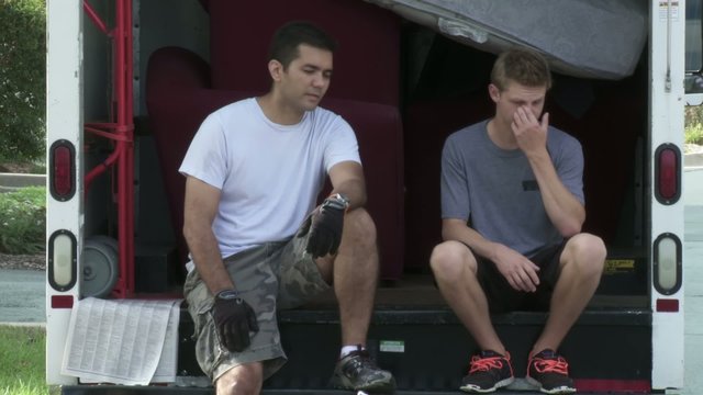 Man And Teenager Sit On Back Of Removal Van, Resting During Move Into An Apartment.