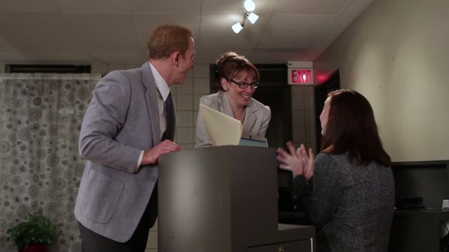 Office workers celebrate a successful deal, then the female boss slaps her male co-worker's backside.  He leaves and she plays it up with a female colleague.  Camera mounted on moving jib arm.