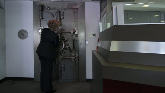 Woman opens a large bank vault door revealing the safe deposit boxes inside.  Wide shot with camera on slider.