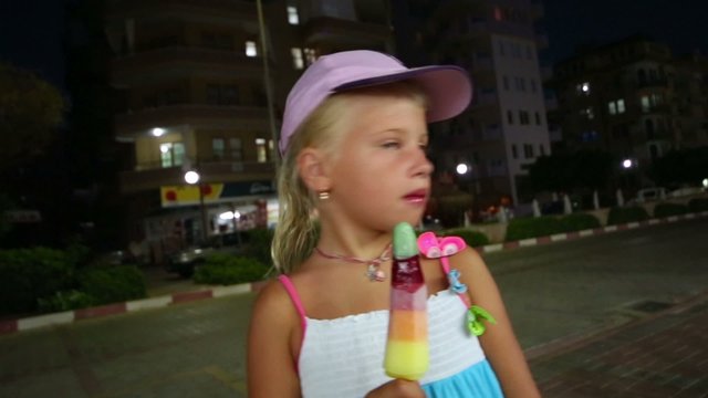 little girl at night eating popsicles on the sidewalk on the background buildings