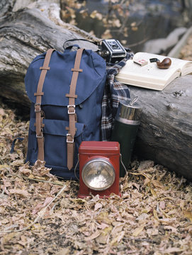 Hiking equipment, a backpack, torch, flask and camera by a tree trunk. A pipe lying on an open book.