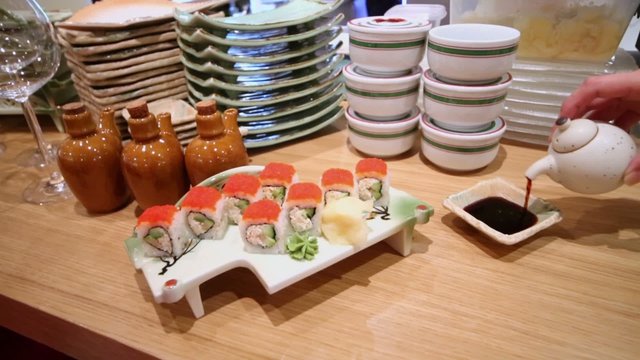 Hand pours sauce to plate on table with rolls at dish