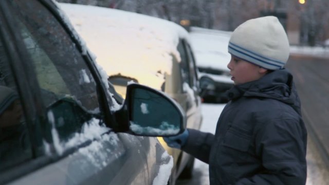 Young boy cleans car from snow at street with tram rides