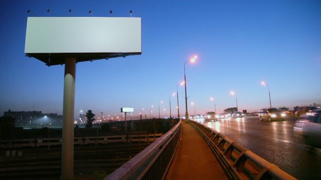 Empty advertising billboard on sidelines of road with traffic near railroad at evening, fast motion time lapse