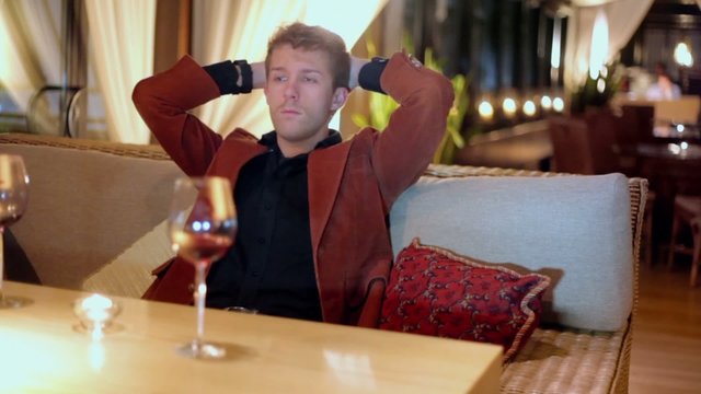 Blond man puts hands behind head during sits at sofa
