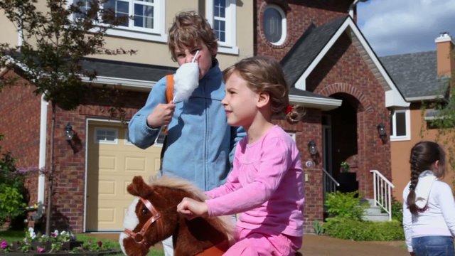 Boy eats candy floss and his little sister sits on toy horse, girl passes near cottage at summer day