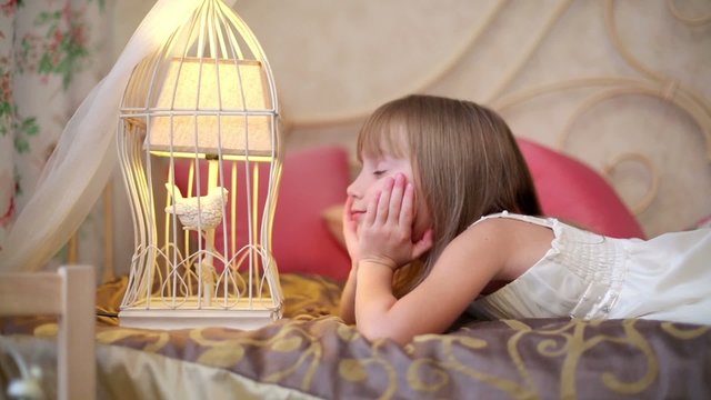 Little girl lying on the bed and looking at a bird in cage