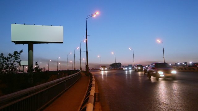 Empty advertising billboard on sidelines of highway with traffic at evening
