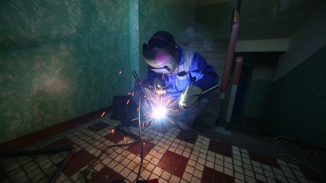 Welder in protective suit and mask welds metal pipes squatting