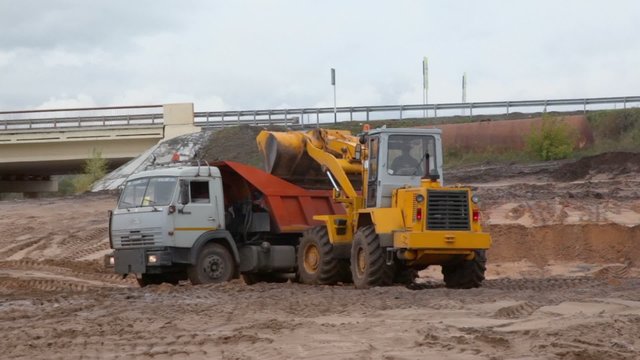 Bulldozer loads truck by sand near road at cloudy day