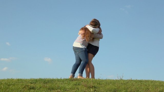 Two young girls embrace and raise each other on grass hill at sunny summer day