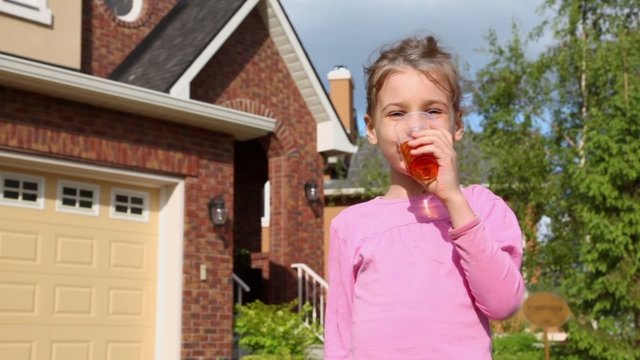 Little girl drinks juce from plastic glass and smiles near cottage at sunny summer day