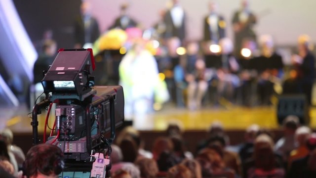 Video camera shoots concert in concert hall with many spectators