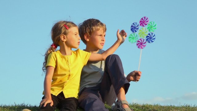 Two kids sit close together talk and play with windmill toy on grass hill at sunny summer day 