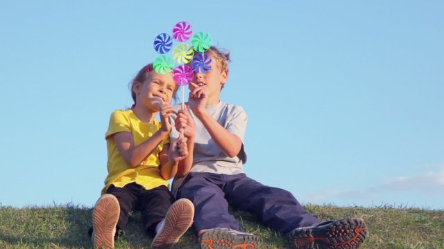 Two kids sit close together on grass hill and play with windmill toy at sunny summer day 