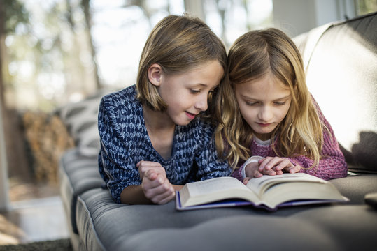 Two blond girls lying on a sofa, looking at a book.