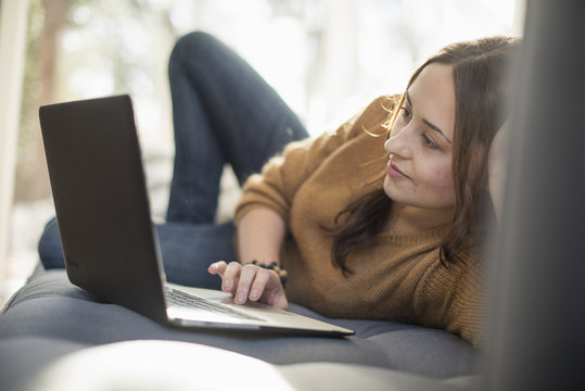 Woman lying on a sofa looking at her laptop, smiling.