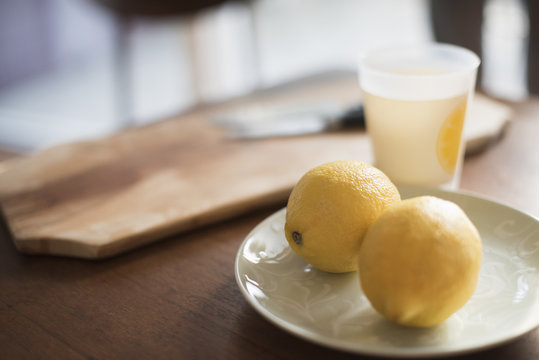A chopping board with knife, a plate with two lemons and a glass on a table.