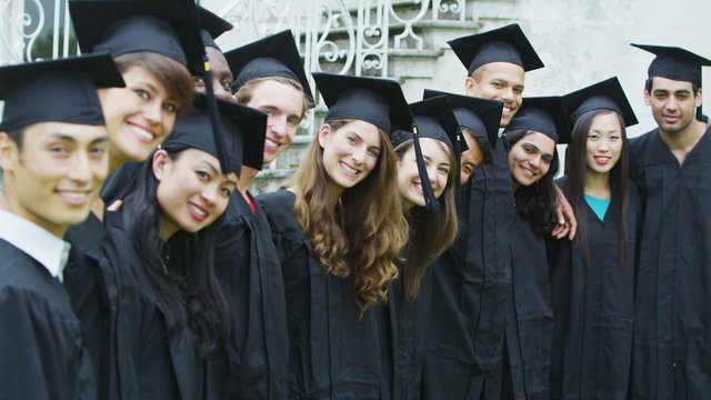 Happy students on graduation day stand in a line & pose for photographs outdoors