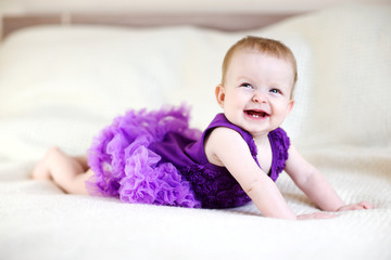 Laughing baby girl in purple dress on white bed