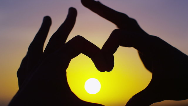 Hands join together to make a heart shape, outdoors on a beach at sunset
