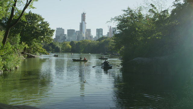 People in rowboats on central park lake with the New York skyline behind them.