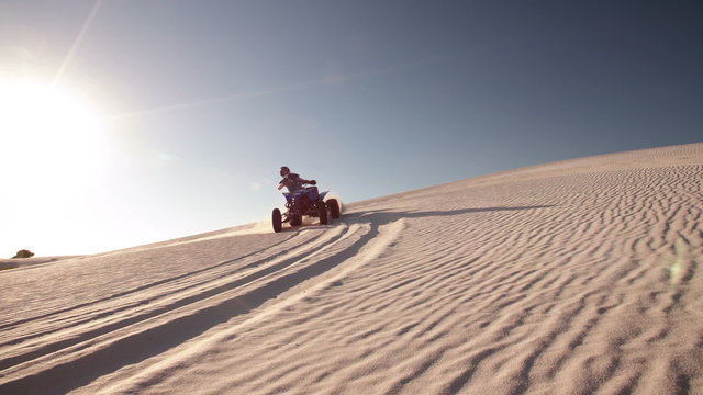Quad racer driving up a sand dune with sun flare