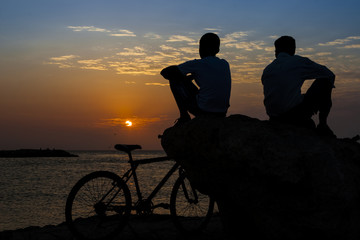 Obraz premium Silueta de dos hombres sentados junto al mar con su bicicleta disfrutando de un hermoso atardecer en Cartagena de Indias en Colombia. Padre e hijo al atardecer