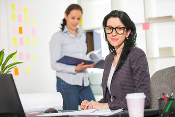 Businesswoman sitting at her desk with colleague on background