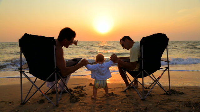 Two Men In Outdoor Chairs And Baby On The Beach At Sunset