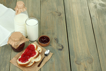Bread with jam, jam in glass jar and jug of milk on wooden table