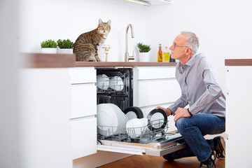 Senior man with cat in the kitchen, empty out the dishwasher
