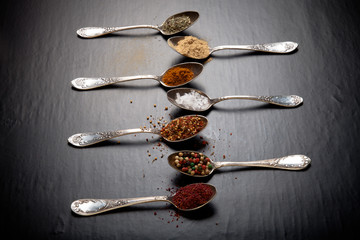 Old metal spoons with different kind of spices on a black backgr