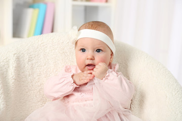Fototapeta na wymiar Cute baby girl in pink dress sitting in arm-chair, on home interior background