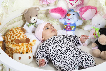 Bebita vestida con una pijama de animal print recostada en su cuna rodeada de animales de peluche
