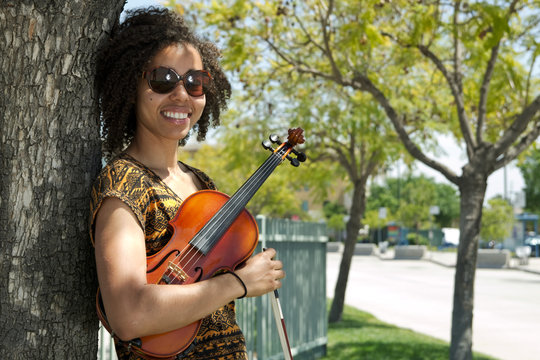 African American woman holding violin and leaning against tree - Powered by Adobe