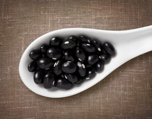 Black beans in white porcelain spoon on sackcloth background