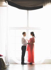 Handsome man and cute pregnant wife standing next to big window
