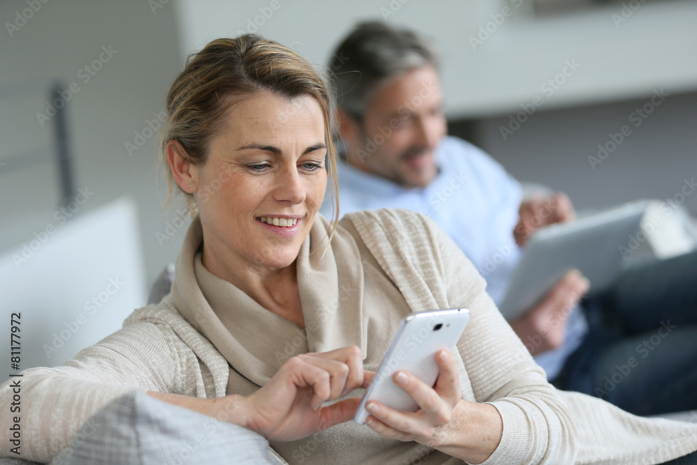 Wall mural Mature woman using smartphone, husband in background