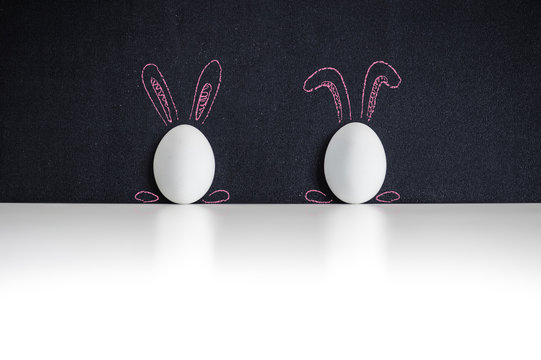Two white easter eggs painted with rabbits ears on a chackboard
