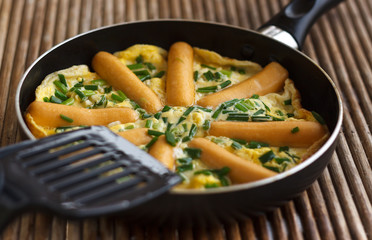 Omelette with sausages and herbs in frying pan on rattan table