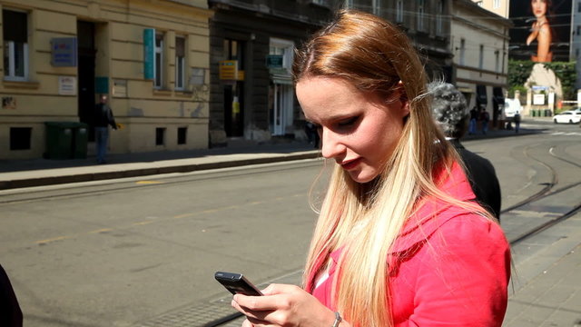 Blond girl in red coat standing on the street, typing on phone